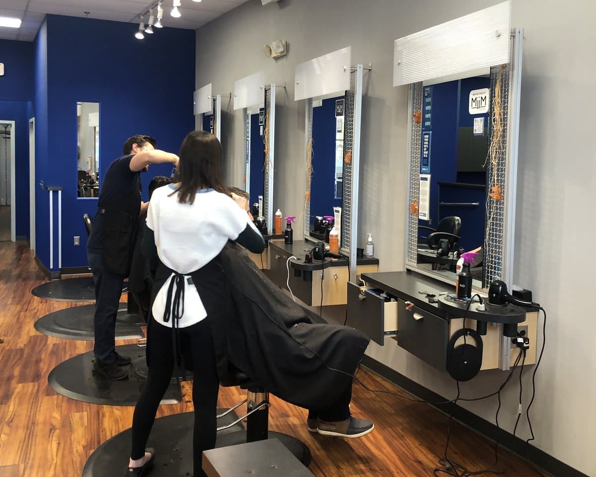 Two MjjM stylists working side by side on caped clients at blue-walled styling stations