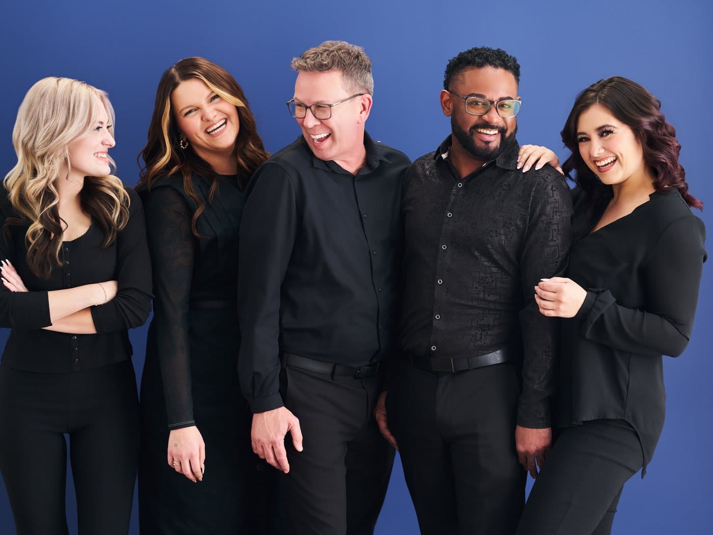 Five Supercuts stylists in all-black laughing together with arms around each other against a blue backdrop